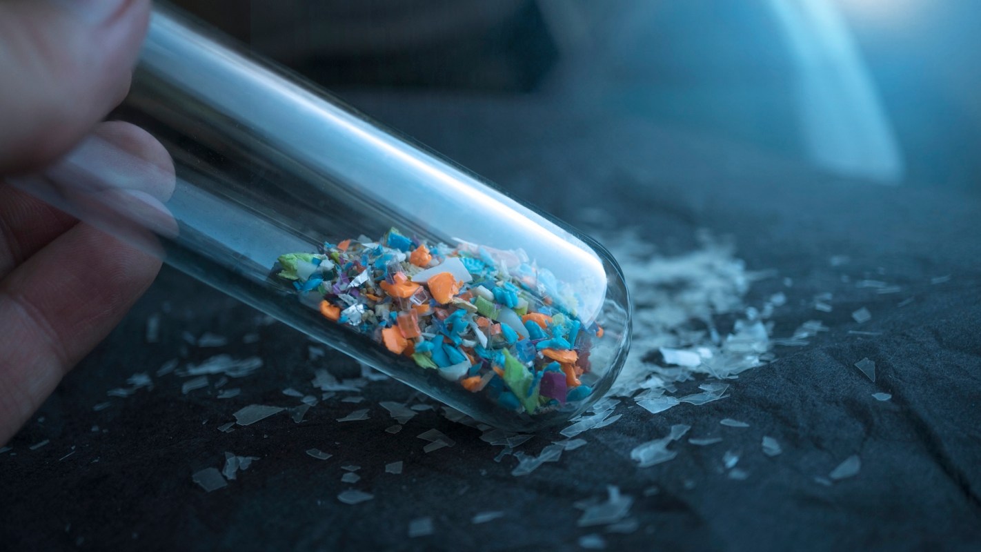 A close-up of a hand holding a glass test tube filled with colorful plastic fragments on a dark surface.