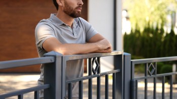 A man leans against a gray metallic gate, contemplating in a sunlit outdoor setting.