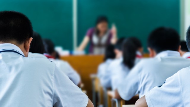 Students in white uniforms sitting at desks, focused on a teacher at the front of a classroom.