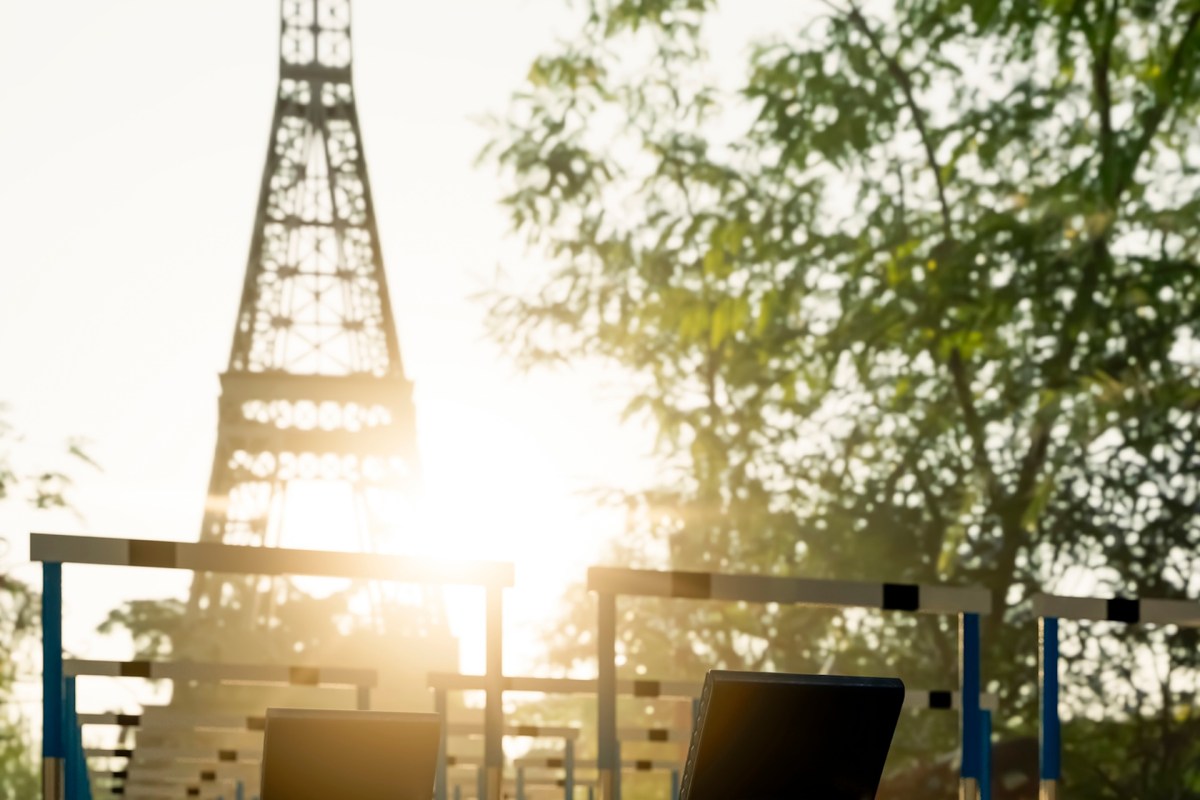 The Eiffel Tower is silhouetted against a bright sunrise, framed by trees and outdoor exercise equipment in the foreground.