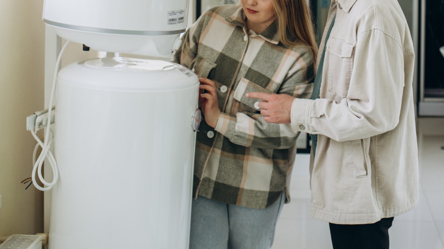 A man and a woman examine a white water heater in a bright indoor space.