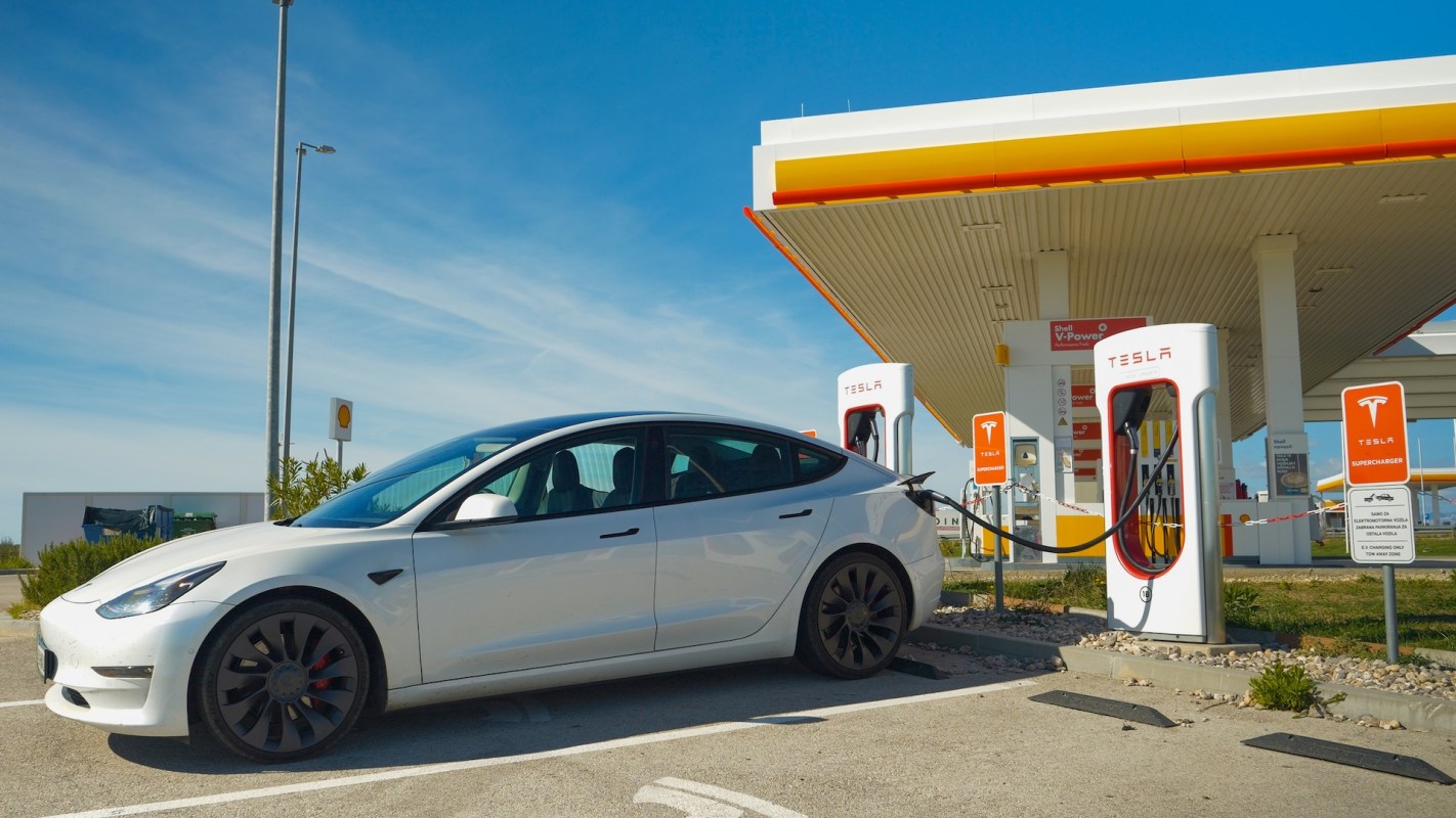A white Tesla Model 3 is charging at a Supercharger station beside a Shell gas station on a sunny day.