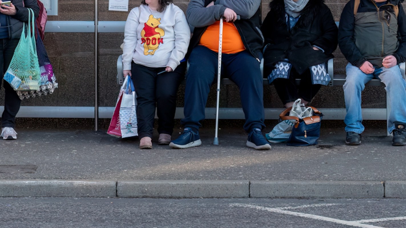 A diverse group of people seated at a bus stop, some holding shopping bags and one using a mobility aid.