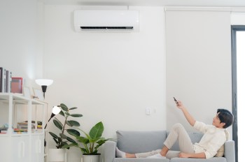 A person relaxing on a couch, using a remote control with a mini-split unit on a wall and houseplants in the background.