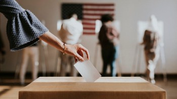 A hand places a ballot into a voting box with people voting in the background and an American flag on the wall.
