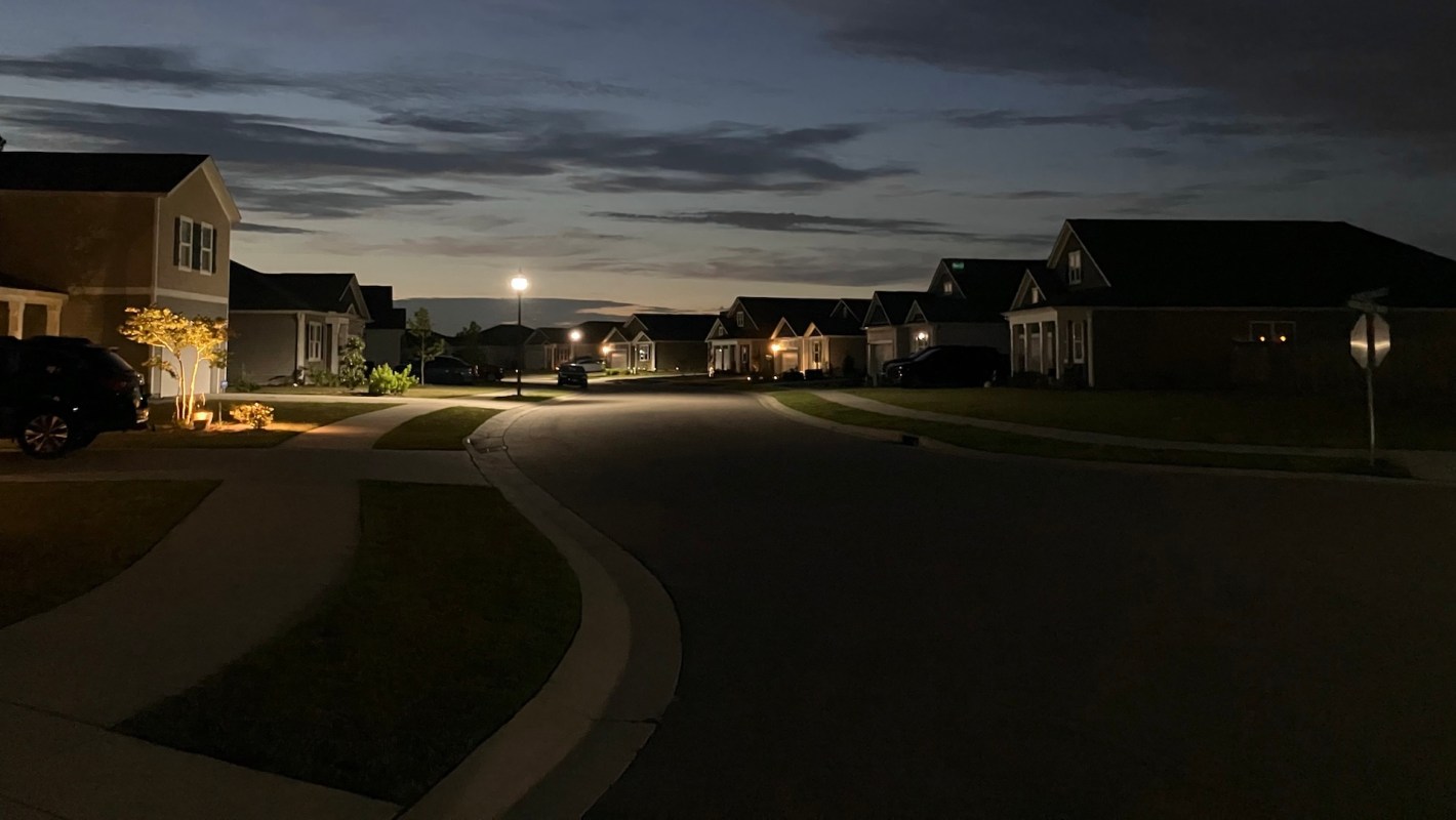 A quiet suburban street at dusk, with softly lit houses and a streetlamp illuminating the sidewalk.