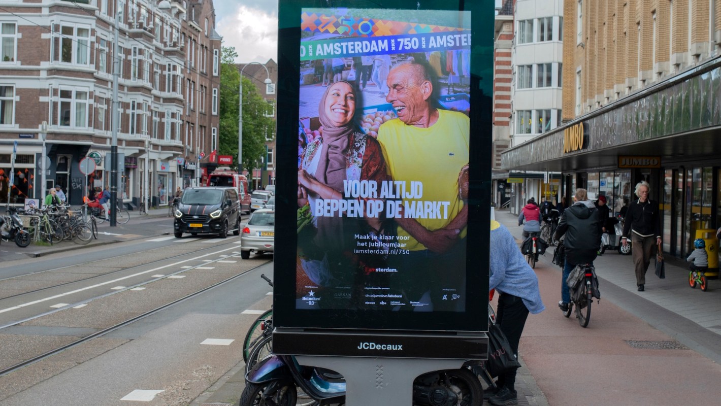A colorful outdoor advertisement features two smiling individuals enjoying a lighthearted moment in Amsterdam.