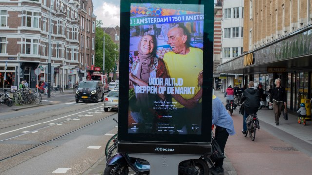 A colorful outdoor advertisement features two smiling individuals enjoying a lighthearted moment in Amsterdam.