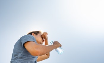 A man wipes his forehead while holding a water bottle under a bright blue sky.