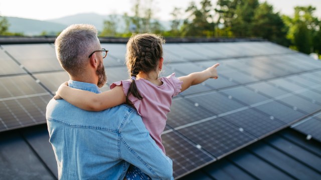 A man holds a young girl on his shoulder as she points at solar panels on a roof.