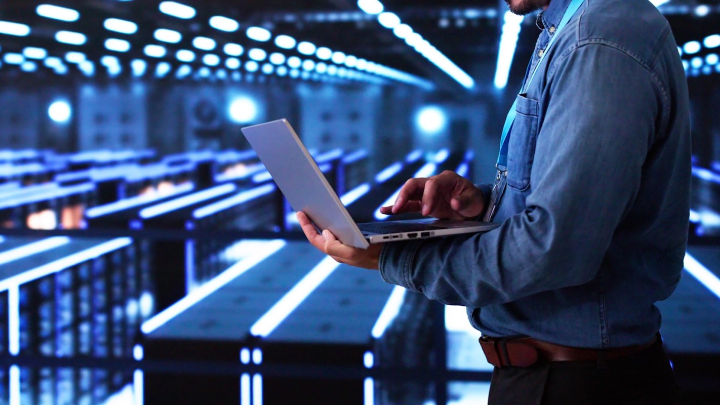 A person in a denim shirt uses a laptop in a data center with glowing rows of servers.
