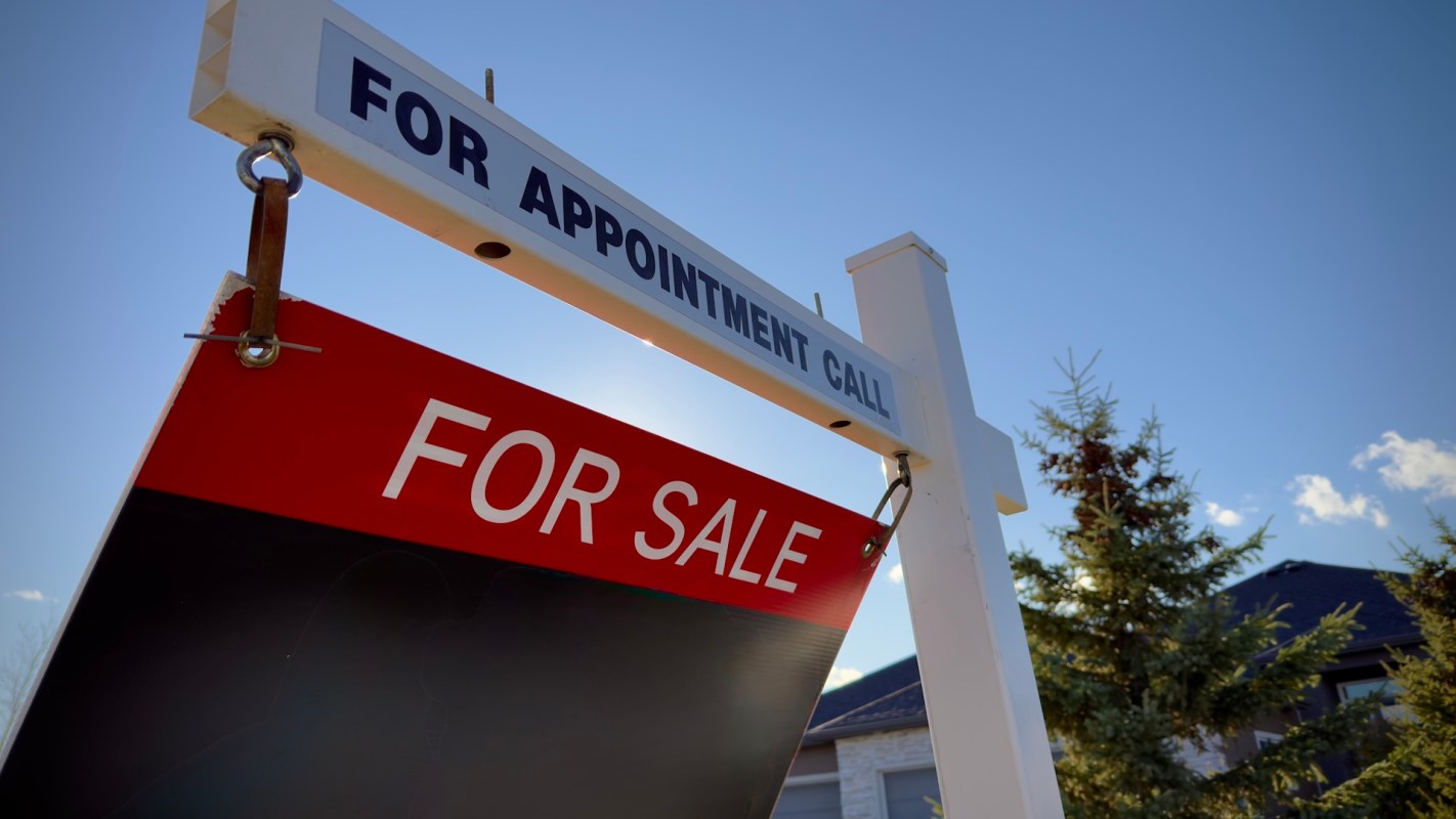 A real estate sign displaying "FOR SALE" against a clear blue sky and trees in the background.