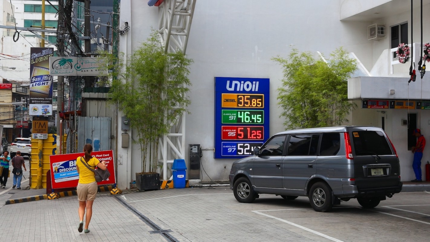 A woman walks toward a gas station with a fuel price board displaying diesel and gasoline rates.