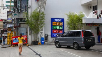 A woman walks toward a gas station with a fuel price board displaying diesel and gasoline rates.