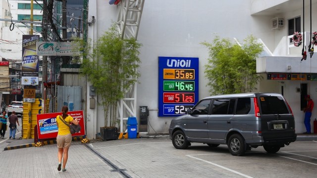 A woman walks toward a gas station with a fuel price board displaying diesel and gasoline rates.