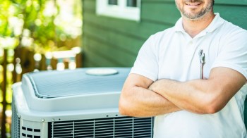 A man in a white shirt stands confidently with arms crossed beside a heat pump unit outside of a home.