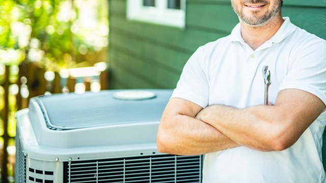A man in a white shirt stands confidently with arms crossed beside a heat pump unit outside of a home.