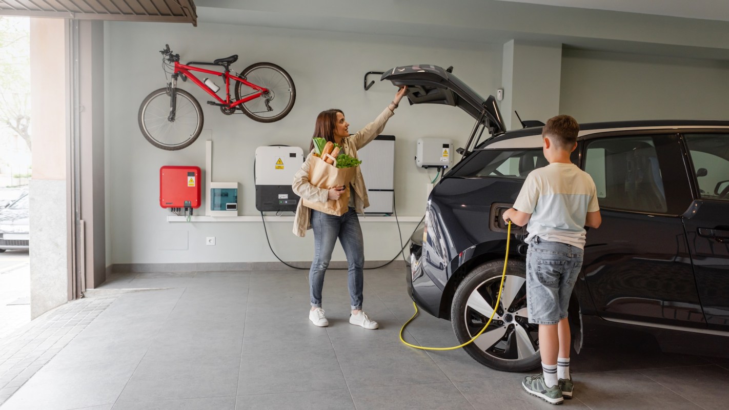 A woman holds groceries while a boy plugs in an electric car in a garage with a bike on the wall.