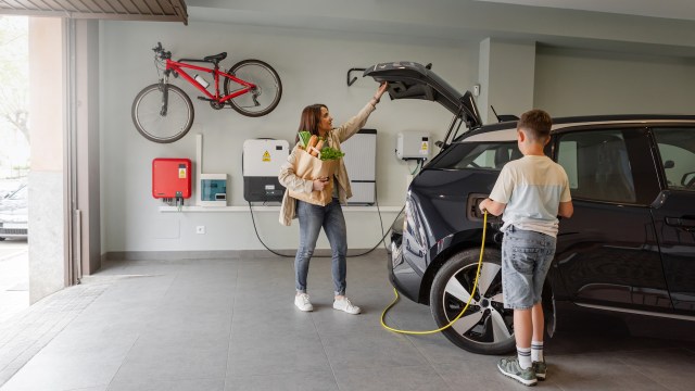 A woman holds groceries while a boy plugs in an electric car in a garage with a bike on the wall.