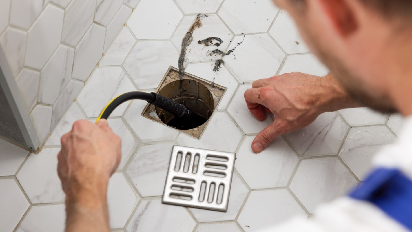 A person using a plumbing snake to clear a drain in a bathroom with hexagonal tiled flooring.