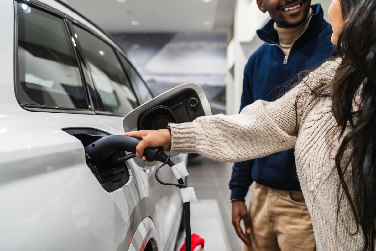 A woman connects a charging cable to an electric vehicle while a man smiles at her.