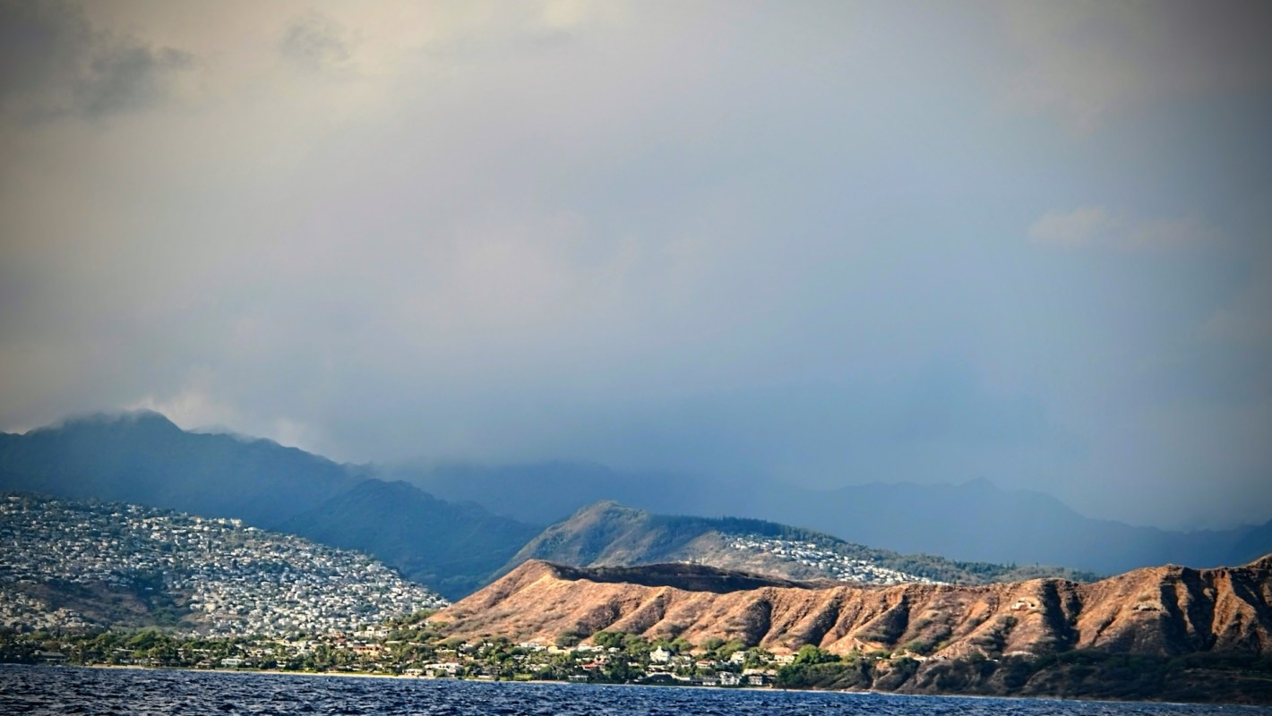 A coastal view featuring a mountainous landscape and a residential area under a cloudy sky.