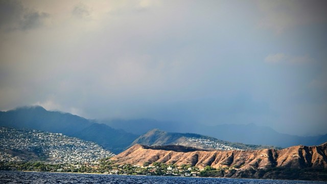 A coastal view featuring a mountainous landscape and a residential area under a cloudy sky.