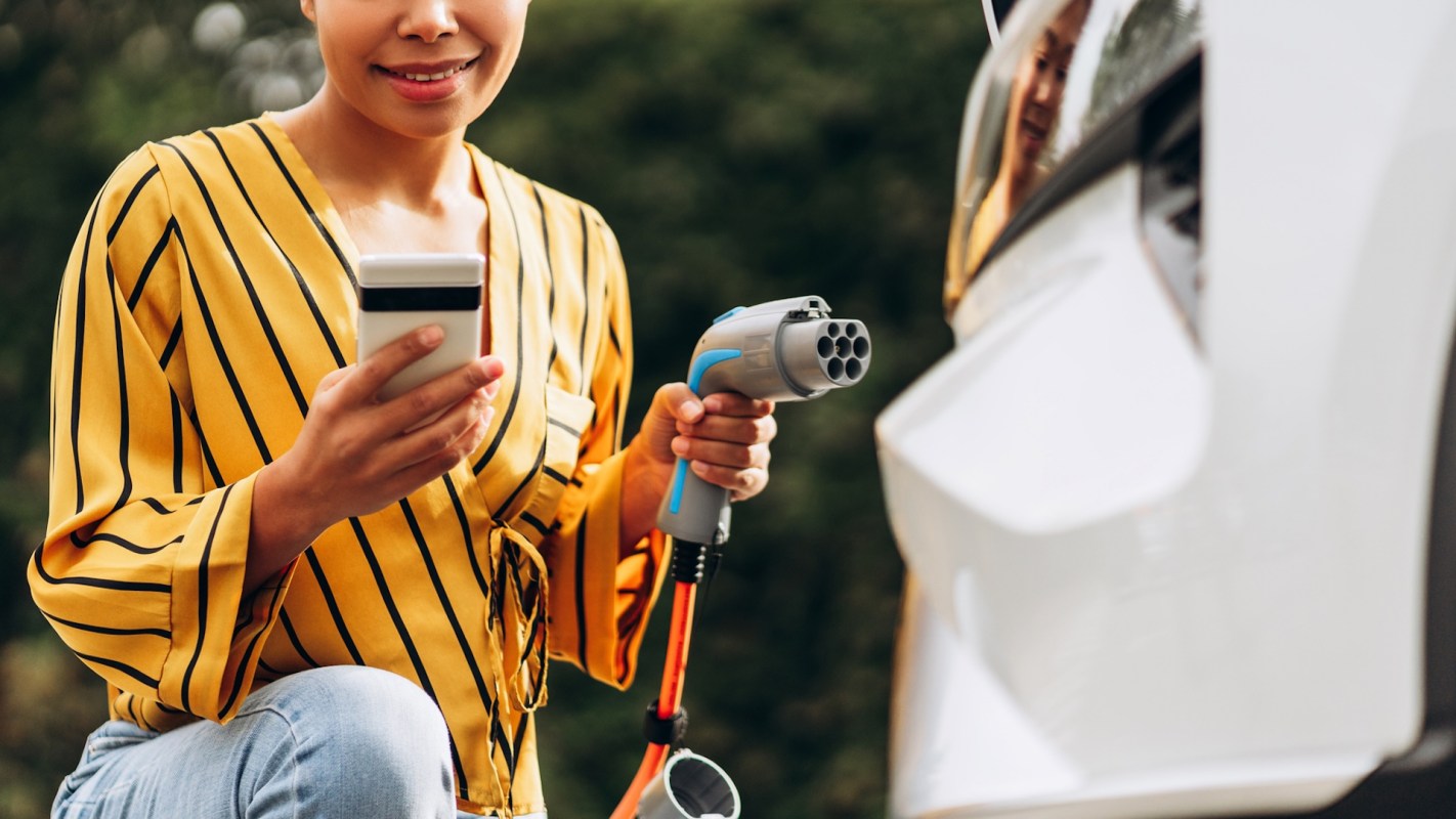 A woman in a striped yellow blouse uses a phone while holding an electric vehicle charger.