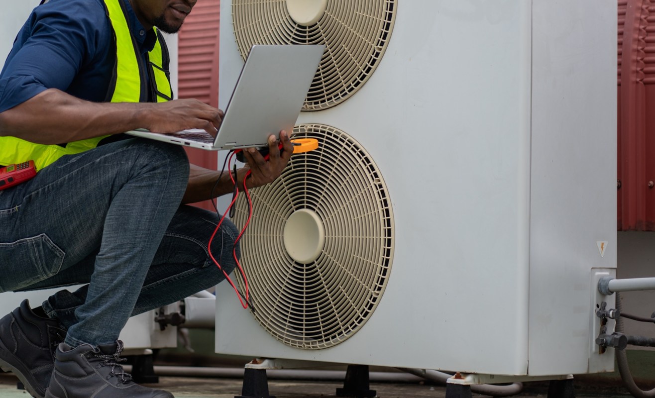 A technician in a safety vest uses a laptop to inspect a heat pump with visible electrical tools.