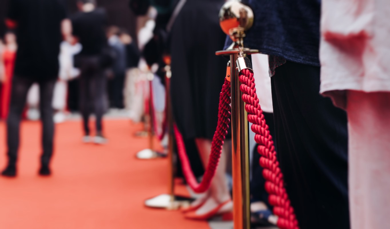 A red carpet event with velvet ropes and blurred figures of attendees in formal attire in the background.