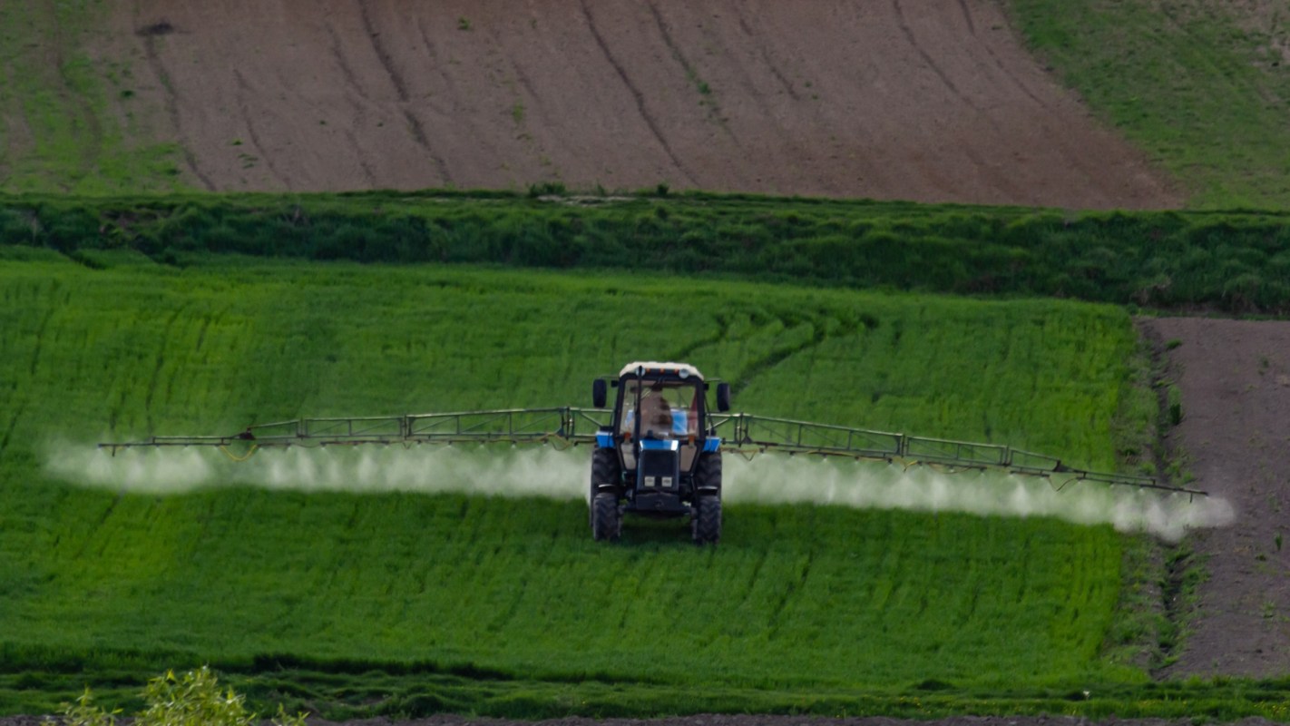 A tractor sprays chemicals over a lush green field with freshly cultivated soil in the background.