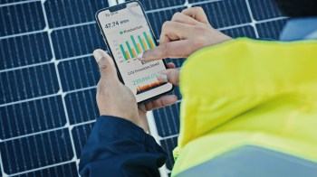 A person in a reflective vest checks a smartphone displaying solar energy production and CO2 emissions data.