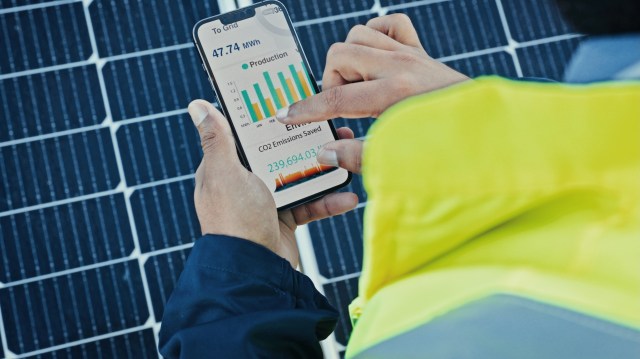A person in a reflective vest checks a smartphone displaying solar energy production and CO2 emissions data.