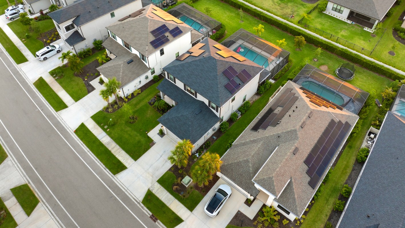 An aerial view of a suburban neighborhood featuring houses with solar panels and green lawns.