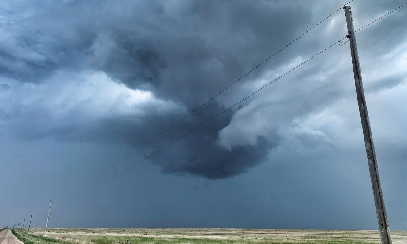 A dramatic storm cloud formation looms over an open landscape with a dirt road and utility poles.