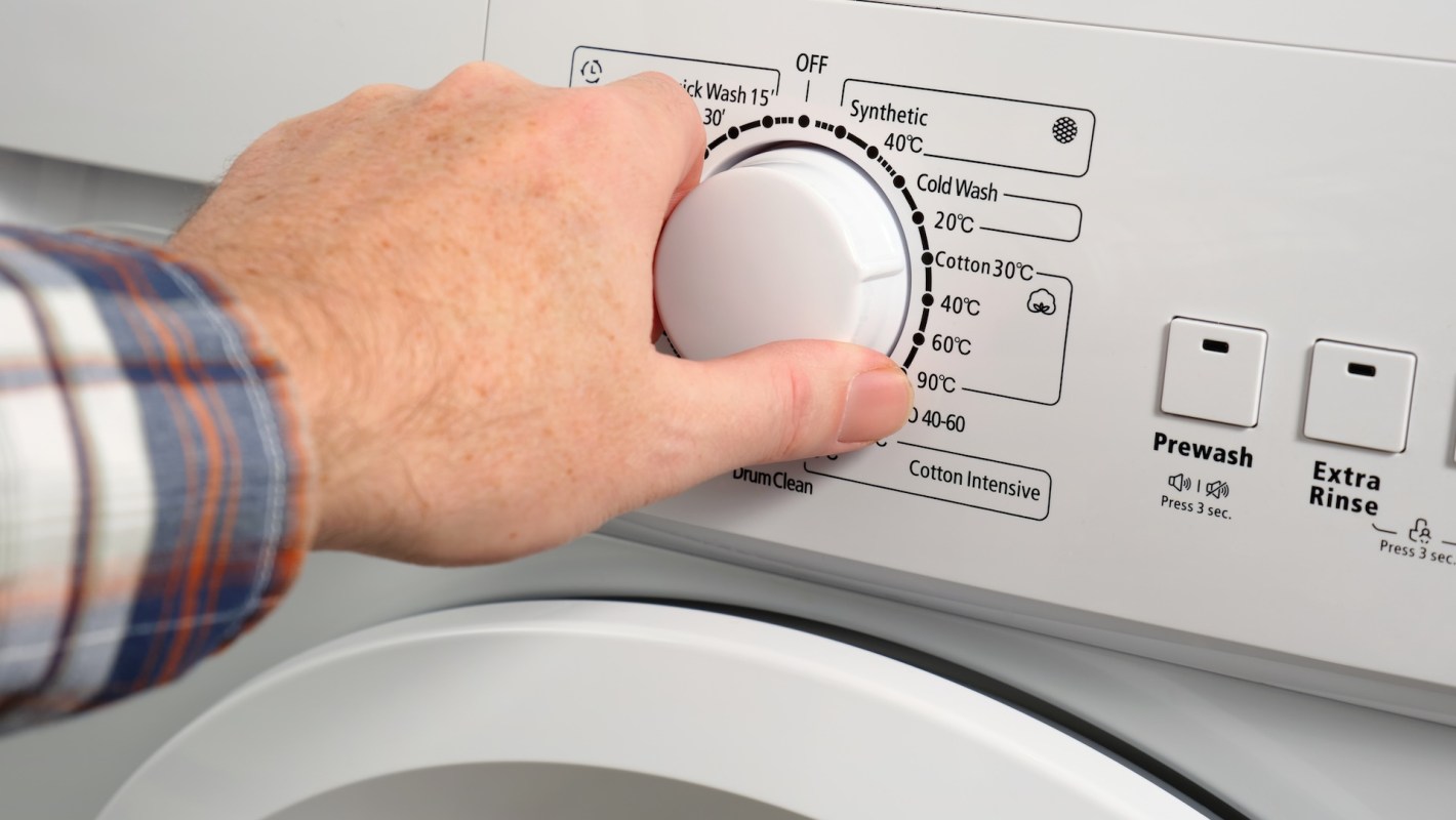 A hand adjusting the temperature dial on a washing machine displaying various wash cycle settings.