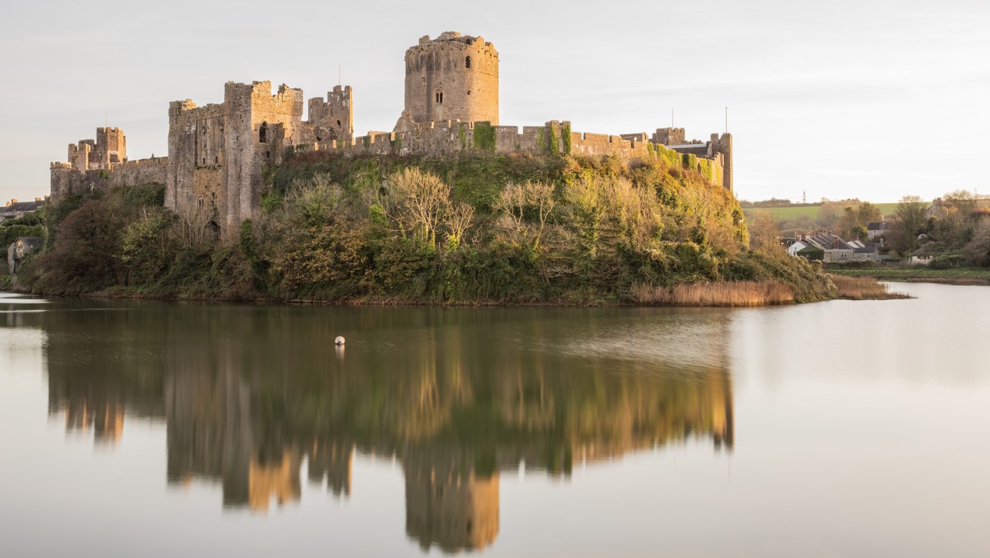 A stone castle sits atop an island surrounded by calm water, reflecting the structure and trees.