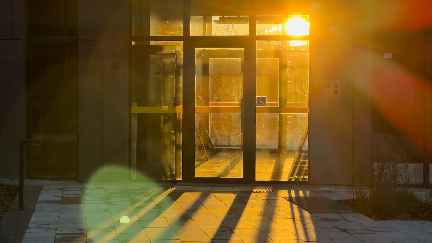 Sunlight streams through glass doors, casting long shadows on the tiled floor of a modern building entrance.
