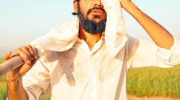 A man wipes sweat from his forehead while holding a tool in a sunny field.