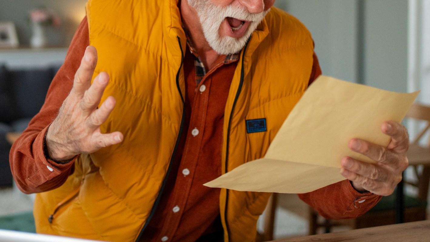 An elderly man in an orange vest reacts with outrage while reading a letter in his hands.
