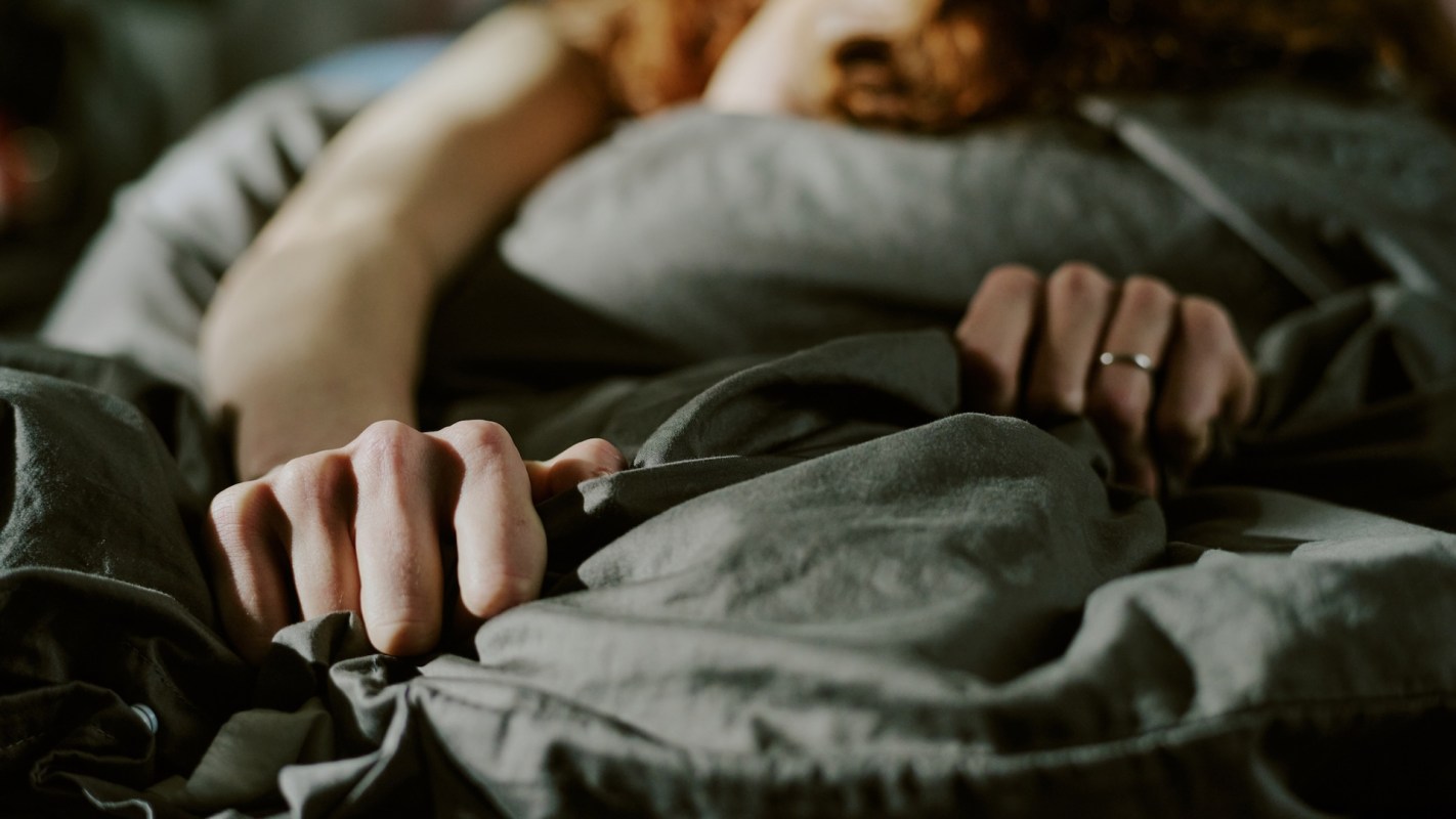 A person lying in bed and gripping gray sheets.