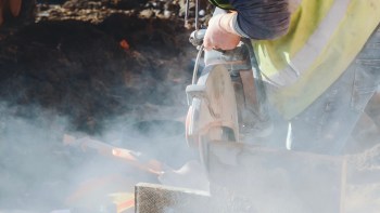 A construction worker using a cutting tool amid dust and debris at a worksite.