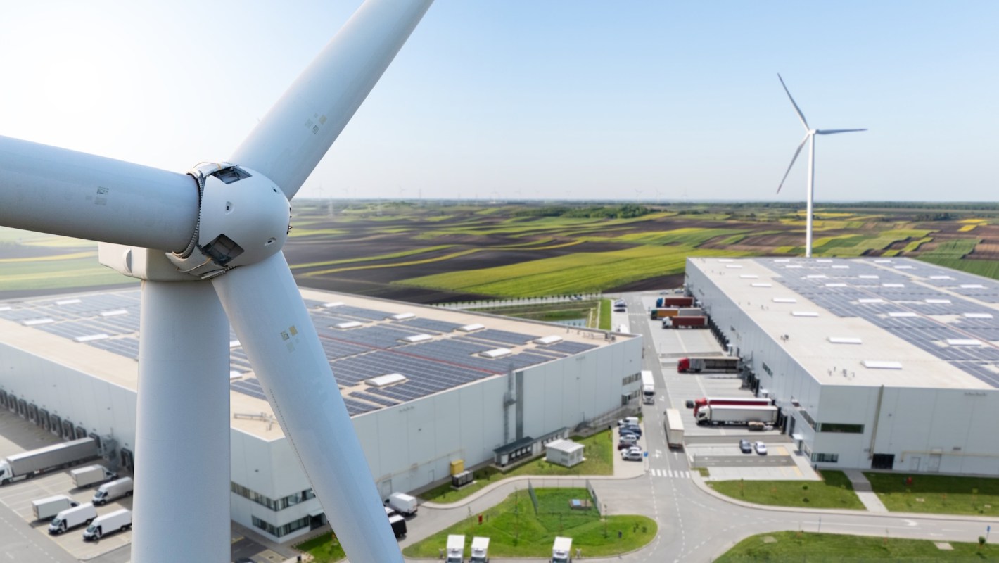 A close-up of a wind turbine with a large warehouse and solar panels in the background, set in a green agricultural landscape.