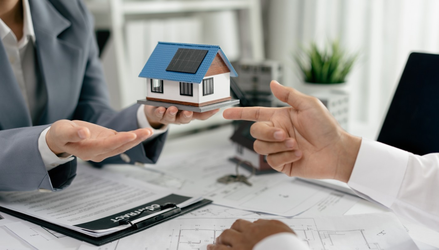 A person is holding a small model house with solar panels while another person reaches out, surrounded by documents.