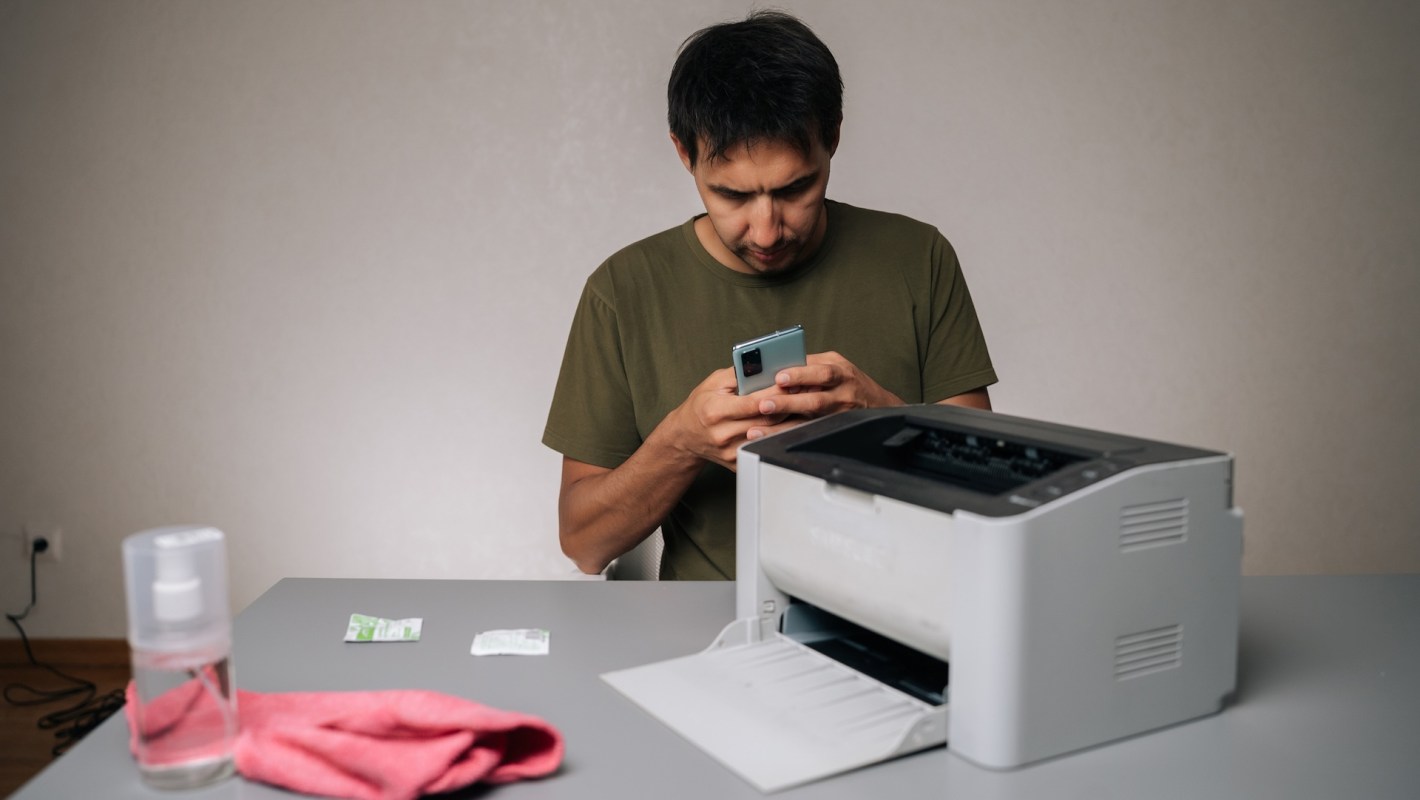 A man using a smartphone while sitting at a table with a printer and cleaning supplies.