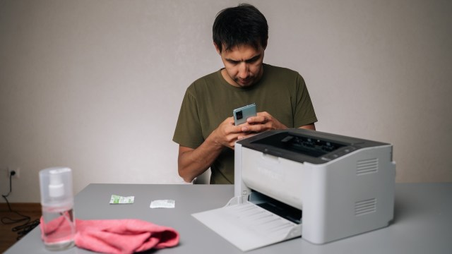 A man using a smartphone while sitting at a table with a printer and cleaning supplies.