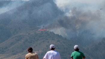 Three young individuals watch a wildfire burning on a mountain, with smoke billowing into the sky.