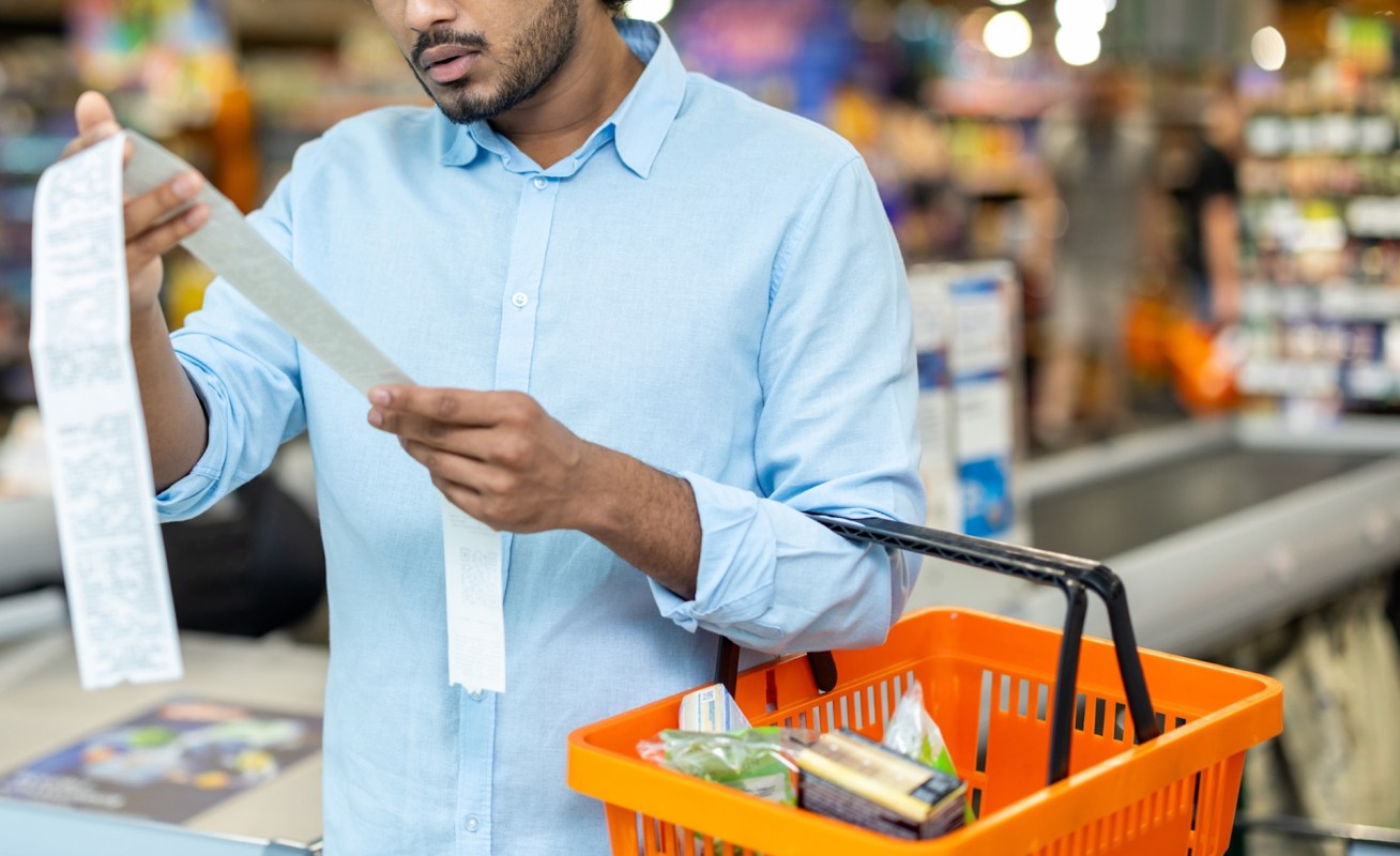 A man in a blue shirt examines a grocery receipt while holding an orange shopping basket in a supermarket.