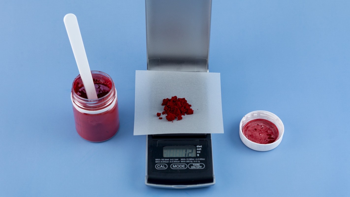 A digital scale weighing red powder on parchment paper beside a jar and a lid of red mixture.