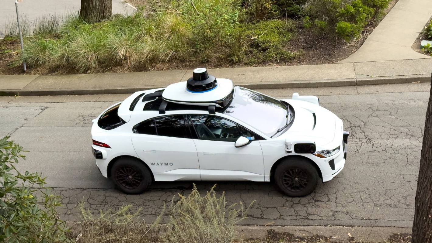 A white Waymo autonomous vehicle with a sensor on top drives along a residential street surrounded by greenery.